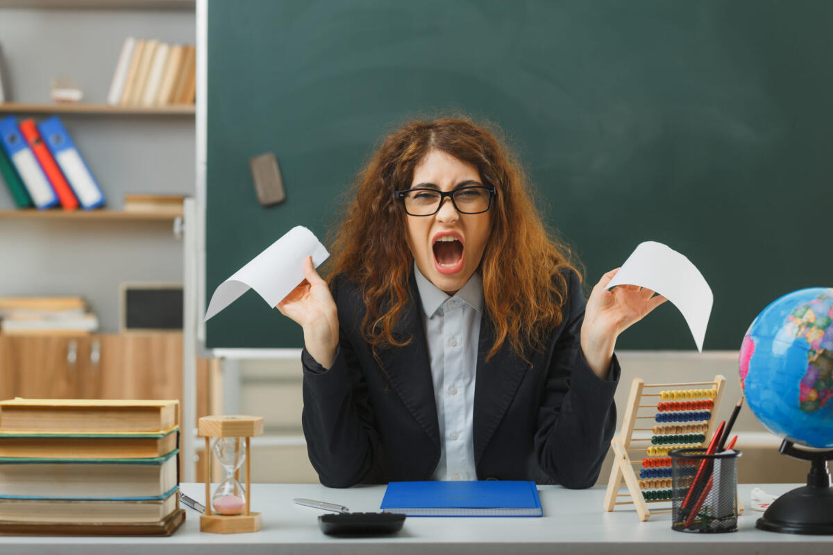 Stipendi docenti e Ata i più bassi della PA, quelli dei precari a picco - angry young female teacher wearing glasses tear paper sitting at desk with school tools in classroom