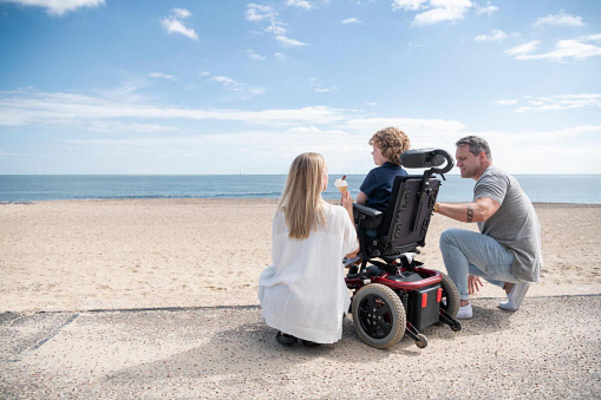 Non lasciamo i genitori soli - Mid adult mother and mature father kneeling by 6 year old child with muscular dystrophy by the beach, enjoying an ice cream while looking at the view. Clacton on Sea.